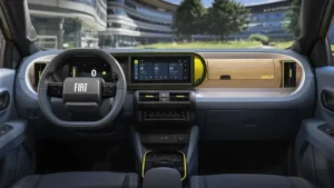 Interior de um carro Fiat com painel horizontal moderno, quadro de instrumentos digital e central multimídia integrada ao centro do painel. O volante multifuncional traz o logotipo FIAT em destaque e design quase quadrado. O acabamento mistura tons escuros, faixa texturizada criando um visual jovem e tecnológico.
