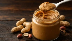 Pote De Pasta De Amendoim Cremosa Com Colher Branca E Amendoins Ao Fundo