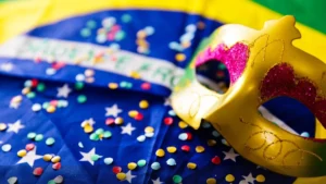 Uma Máscara De Carnaval Dourada, Com Detalhes Em Rosa E Brilho, Apoiada Sobre Uma Bandeira Do Brasil Coberta Por Confetes Coloridos. A Composição Transmite O Clima Festivo Do Carnaval Brasileiro, Unindo Elementos De Celebração, Cultura Nacional E Alegria Típica Da Folia