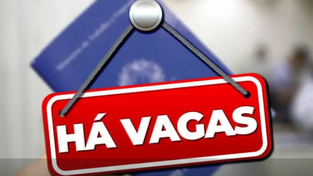 Imagem Mostra Uma Placa Vermelha Com A Frase “Há Vagas”, Pendurada Por Fios, Simbolizando Oportunidades De Emprego Disponíveis. Ao Fundo, Desfocado, Aparece Uma Carteira De Trabalho, Reforçando O Tema De Contratação E Mercado De Trabalho