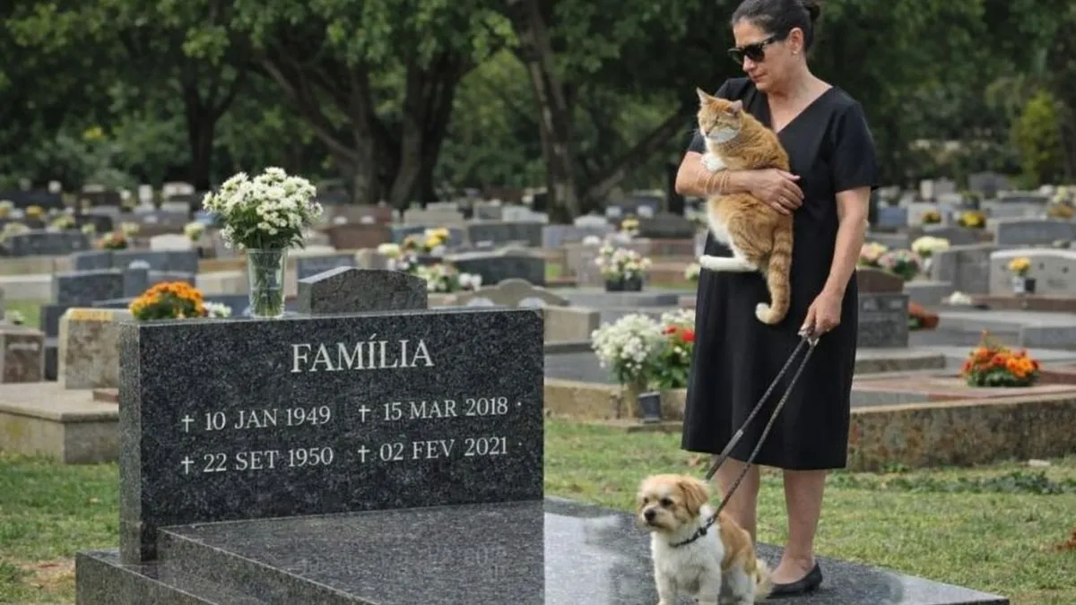 Mulher vestida de preto segura um gato no colo e conduz um cachorro com coleira ao lado de jazigo familiar em cemitério durante o dia