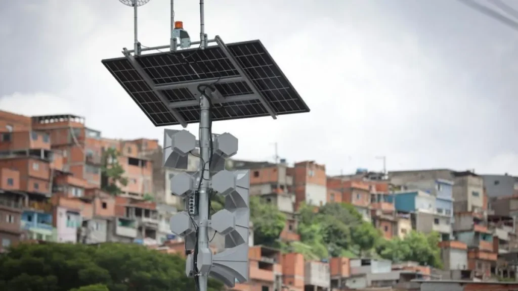 Sirene De Alerta Para Deslizamentos Instalada Em Área Urbana Com Moradias Em Encosta Ao Fundo.