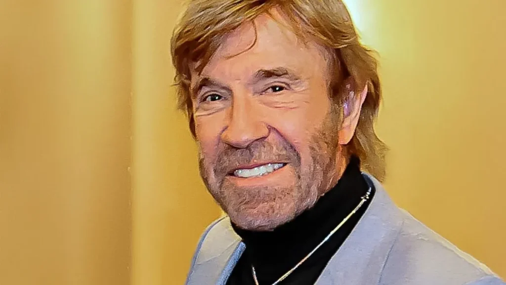 Chuck Norris Sorrindo Em Retrato Recente, Usando Blazer Claro E Gola Alta Preta, Com Fundo Neutro Iluminado