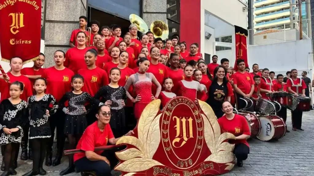 Grupo De Jovens E Crianças Uniformizados Em Vermelho Durante Apresentação Da Fanfarra Walter Weiszflog Em Caieiras, Com Instrumentos Musicais E Estandarte Ao Centro