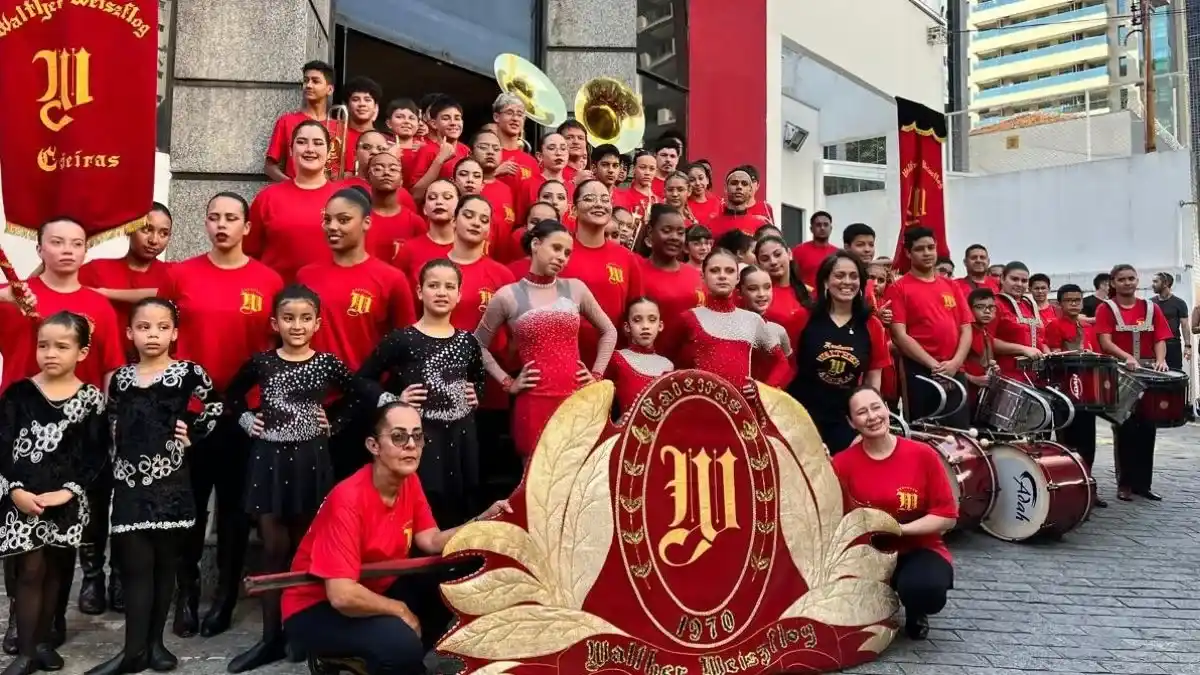 Grupo de jovens e crianças uniformizados em vermelho durante apresentação da fanfarra Walter Weiszflog em Caieiras, com instrumentos musicais e estandarte ao centro