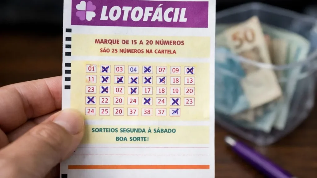 Bilhete Da Lotofácil Sendo Segurado Na Mão Com Números Marcados, Ao Fundo Notas De Real E Caneta Sobre Mesa De Madeira