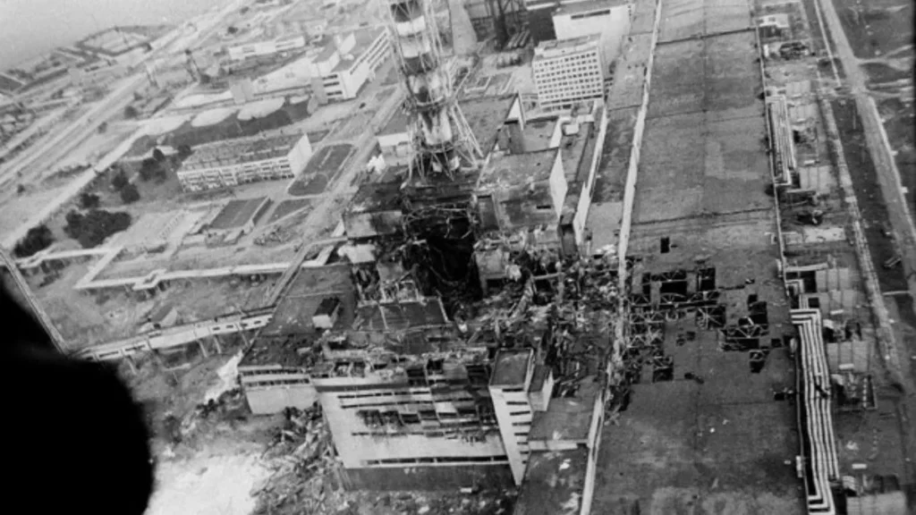 Imagem Em Preto E Branco Mostra O Reator Destruído Da Usina De Chernobyl Após A Explosão De 1986, Com Estrutura Aberta, Destroços Visíveis E Áreas Industriais Ao Redor