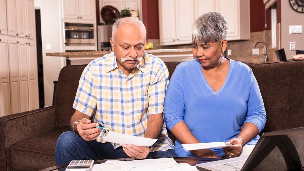Casal De Idosos Analisando Contas E Documentos Financeiros Em Casa, Com Calculadora E Notebook Sobre A Mesa
