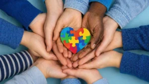Mãos De Diferentes Pessoas, Incluindo Duas Negras E Várias Brancas, Unidas Segurando Um Coração Colorido Com Símbolo Do Autismo Em Destaque