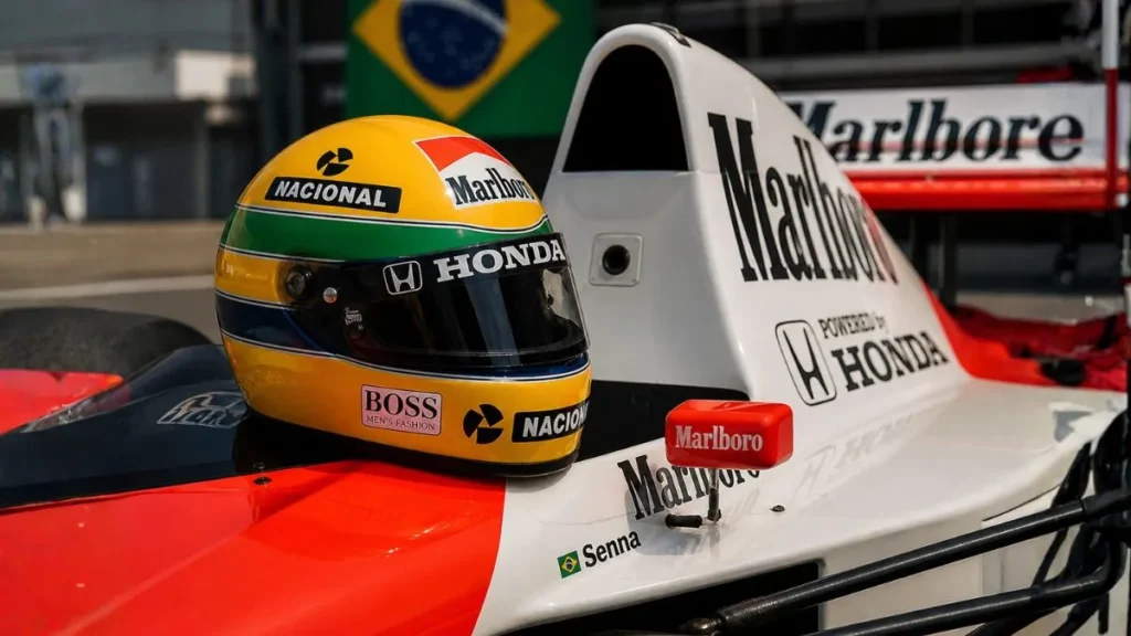Capacete Amarelo Icônico De Ayrton Senna Apoiado Sobre Carro De Fórmula 1 Clássico Com Detalhes Da Mclaren E Bandeira Do Brasil Ao Fundo.