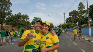 Casal De Torcedores Usando Camisetas Da Campanha “Caieiras Na Torcida Pelo Hexa” E Bonés Do Brasil Posa Com Copos Personalizados Na Avenida Dos Estudantes, Em Caieiras, Durante Ação Em Clima De Copa.