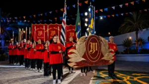 Fanfarra De Caieiras Se Apresenta Na 71ª Festa De Santo Antônio De Caieiras, Com Integrantes Em Uniforme Vermelho, Bandeiras E Estandarte Em Destaque Durante Apresentação Noturna.