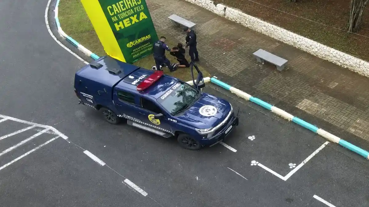 Viatura Da Guarda Civil Municipal De Caieiras Parada Em Via Pública Enquanto Agentes Abordam Um Homem Ao Lado De Um Banner Com A Frase “Caieiras Na Torcida Pelo Hexa”.