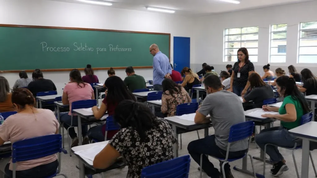 Candidatos Fazem Prova Em Sala De Aula Durante Processo Seletivo Para Professores, Acompanhados Por Dois Examinadores Que Circulam Entre As Carteiras.