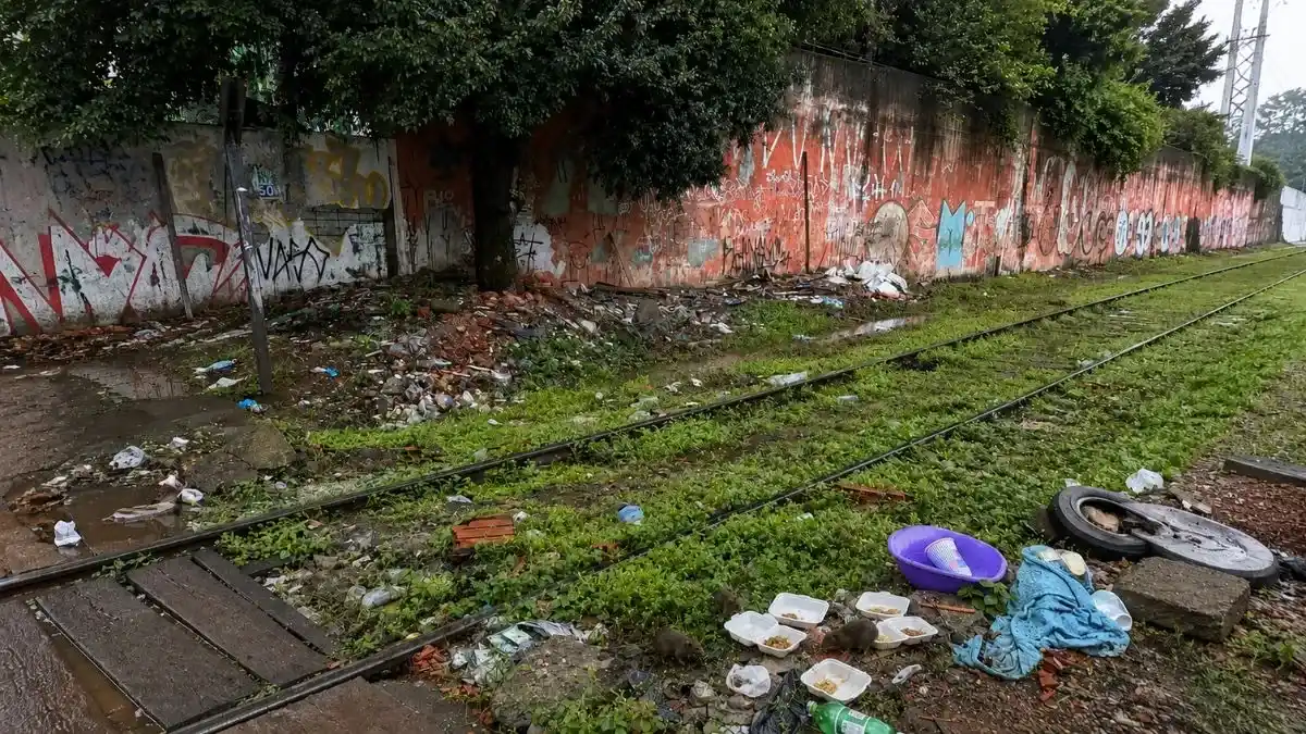 Trilhos de trem com lixo acumulado, marmitas descartadas, garrafas PET, mato alto e ratos próximos aos resíduos em área ferroviária degradada.