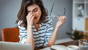 Mulher Cansada Em Frente Ao Notebook Segura Os Óculos E Pressiona A Região Dos Olhos Durante Um Momento De Sobrecarga Mental.
