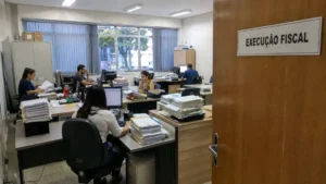 Sala Administrativa Com Placa “Execução Fiscal” Na Porta Aberta, Mostrando Mesas Com Computadores, Servidores Trabalhando E Pilhas De Documentos Sobre Cobrança Pública.