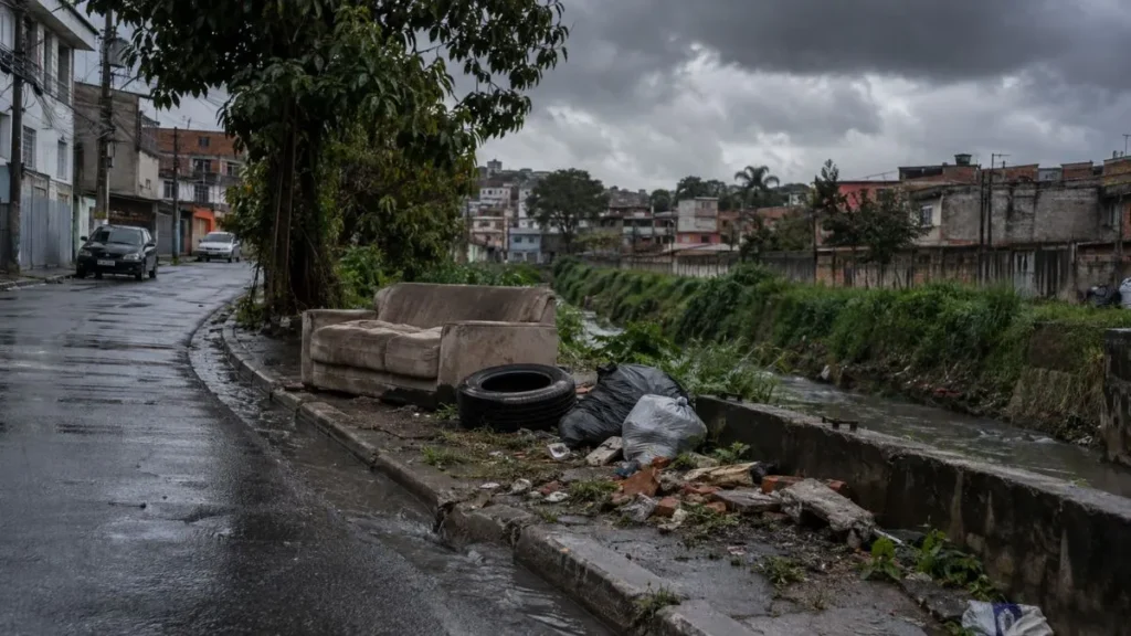 Sofá, pneu, sacos e entulho aparecem descartados irregularmente próximos a uma rua e a um córrego