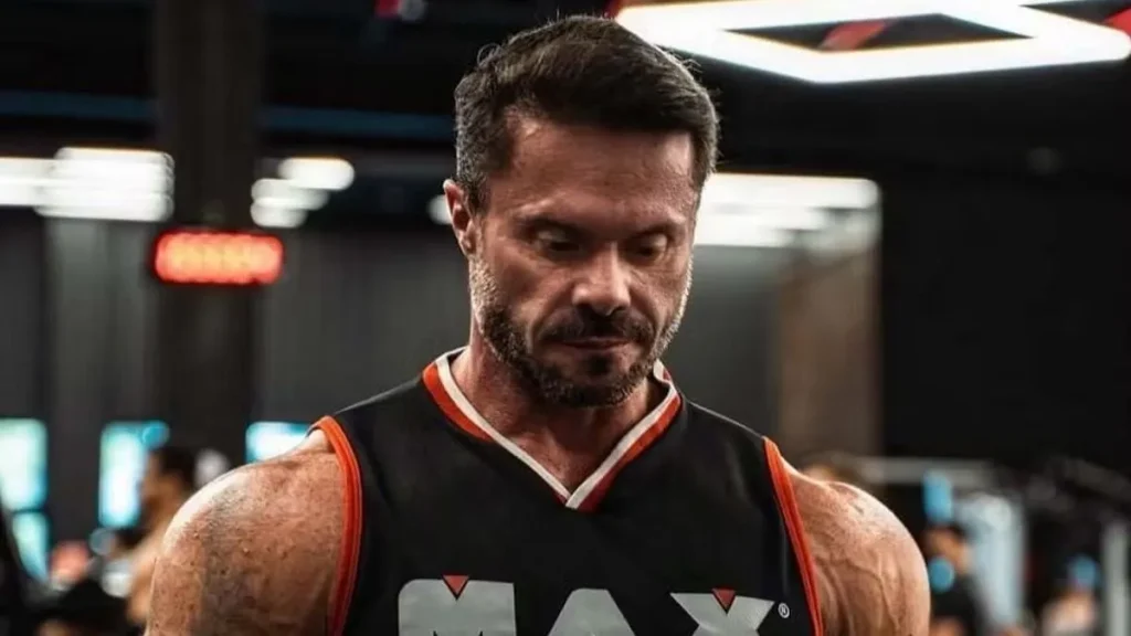 Cariani musculoso, de cabelos curtos e barba, usando camiseta preta com detalhes laranja, fotografado em uma academia, com o olhar direcionado para baixo.