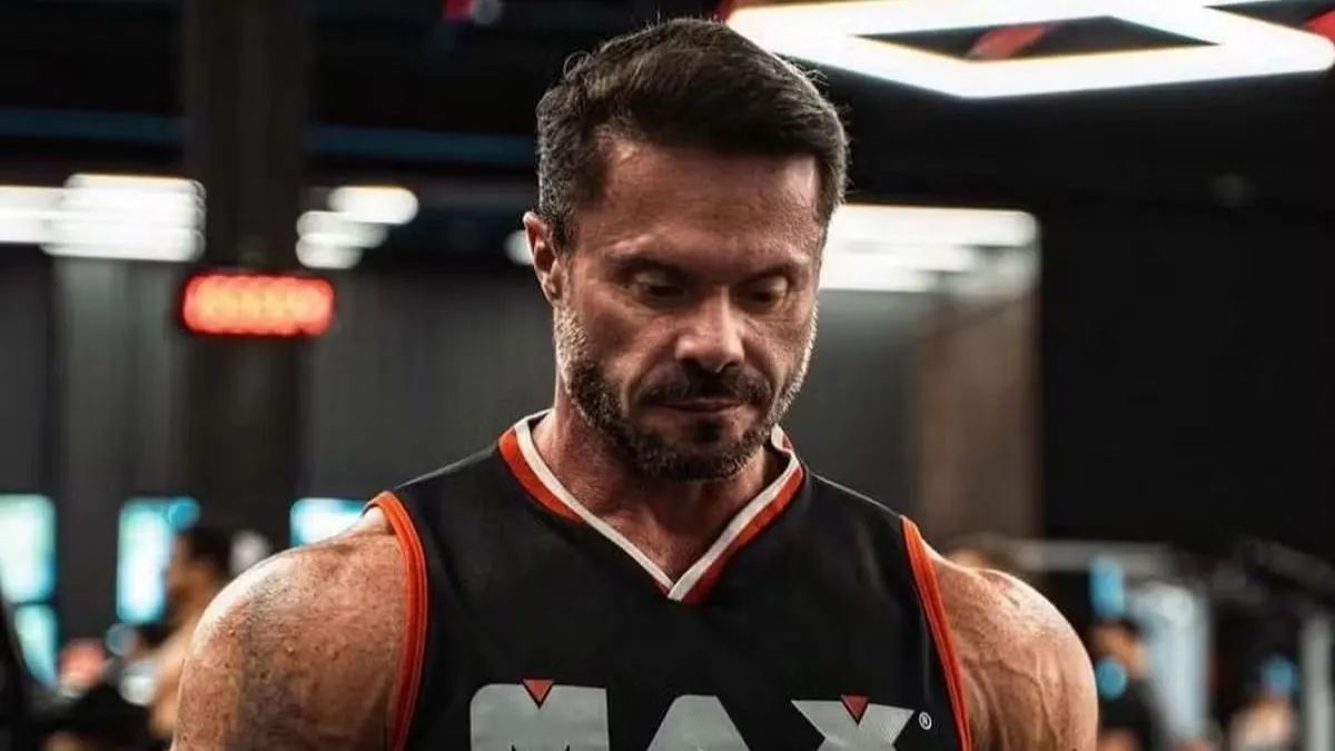 Cariani musculoso, de cabelos curtos e barba, usando camiseta preta com detalhes laranja, fotografado em uma academia, com o olhar direcionado para baixo.
