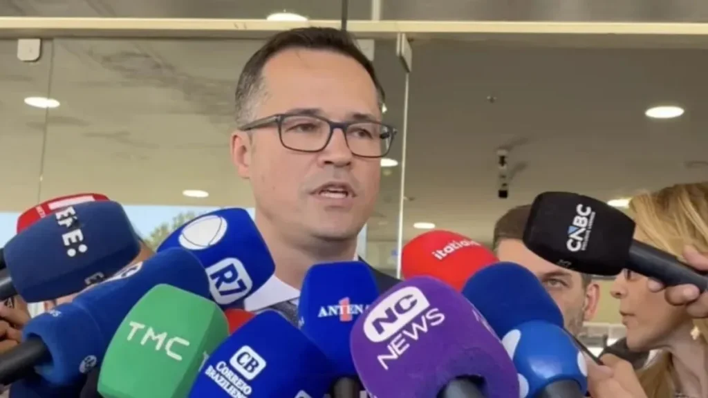 Deltan Dallagnol, de terno e óculos, concede entrevista coletiva, cercado por microfones de diferentes veículos de comunicação.