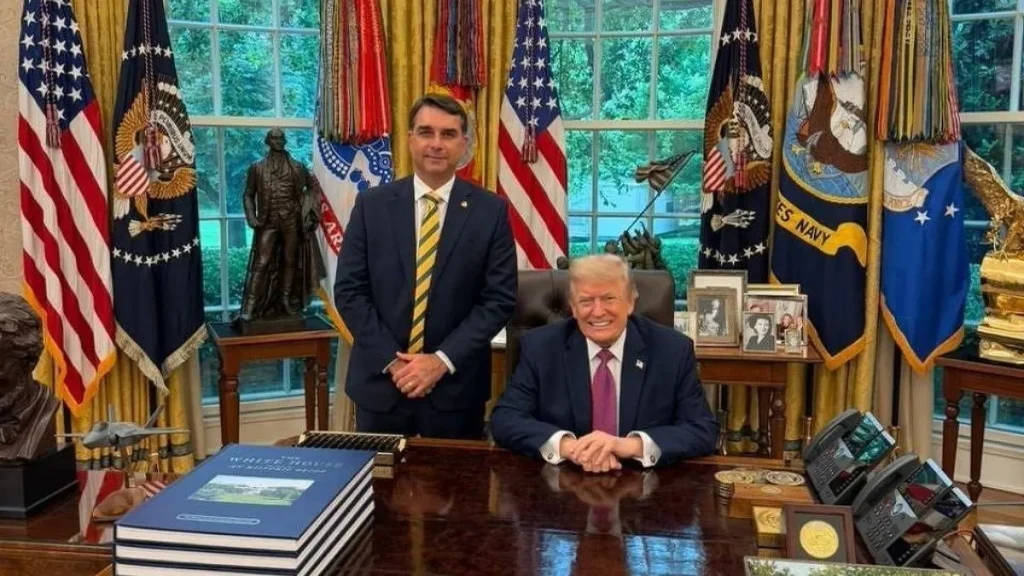 Flávio Bolsonaro pode receber asilo político de Trump nos Estados Unidos? 4 Donald Trump sentado à mesa do Salão Oval da Casa Branca, ao lado de um homem de terno e gravata com as cores do Brasil, diante de bandeiras americanas.