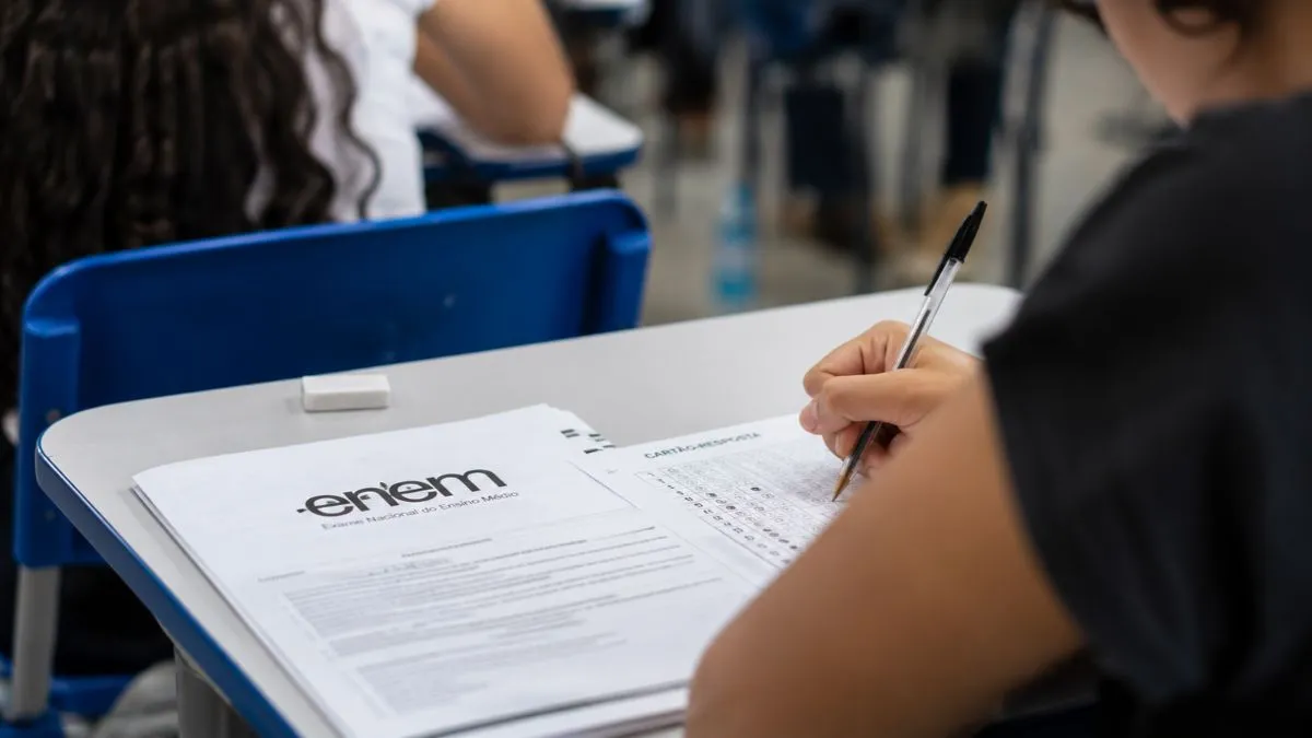 Estudante preenche o cartão-resposta do Enem com caneta preta de corpo transparente, enquanto o caderno de questões está sobre a carteira escolar