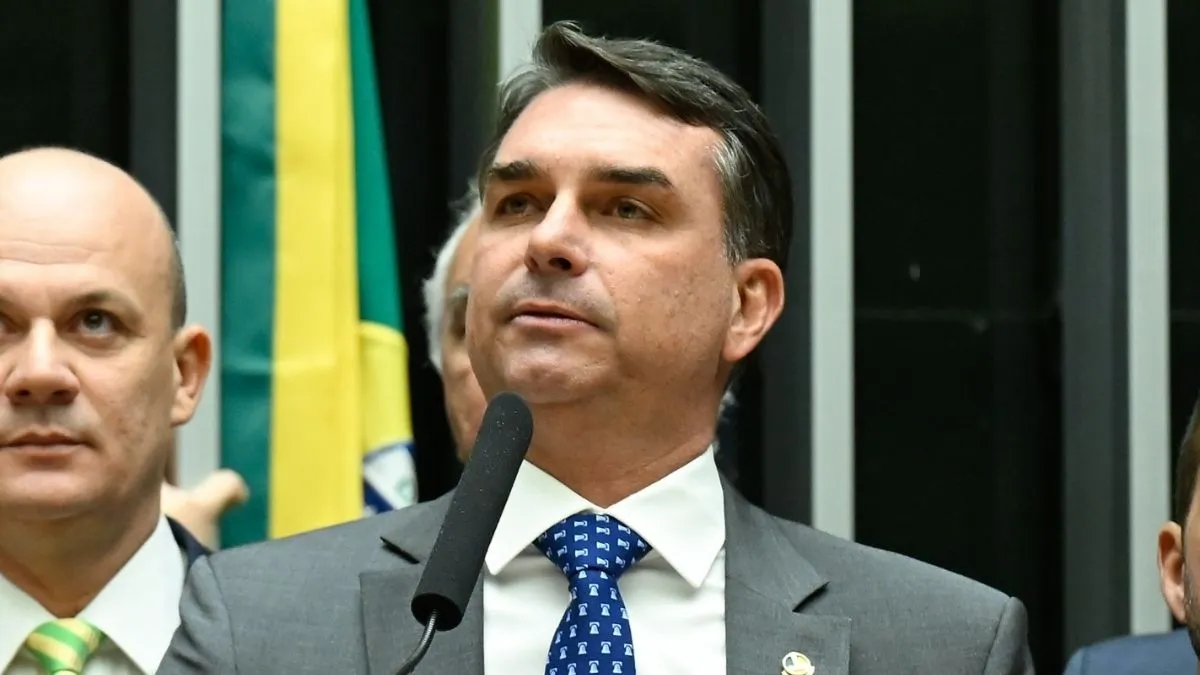 Flávio Bolsonaro (PL-RJ) discursa ao microfone, com a bandeira brasileira ao fundo. Senador teria deixado o Brasil rumo aos Estados Unidos após descumprir ordem do STF.