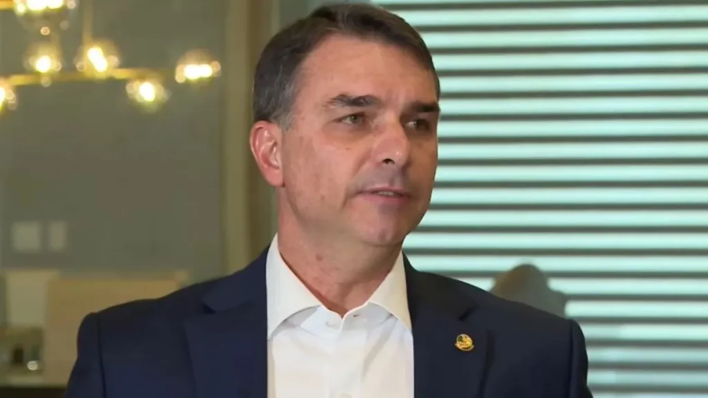 Como Flávio Bolsonaro saiu do Brasil com os passaportes cancelados? 3 Flávio Bolsonaro aparece de terno azul-marinho e camisa branca durante entrevista, em ambiente interno, com persianas e iluminação ao fundo.