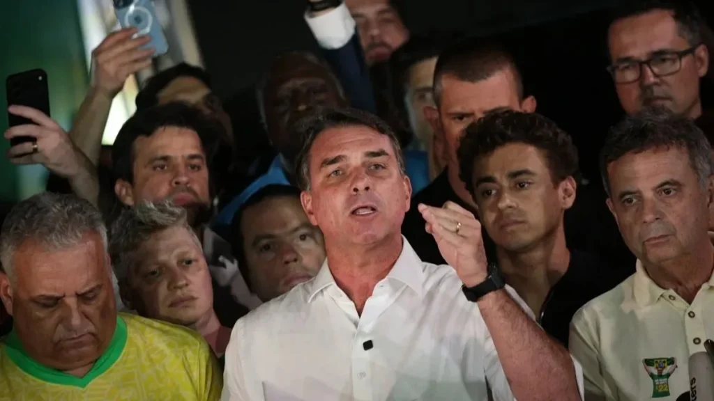 Flávio Bolsonaro, de camisa branca, discursa diante de apoiadores após derrota eleitoral. Senador descumpriu ordem do STF para entregar passaportes, levantando dúvidas sobre a possibilidade de prisão.