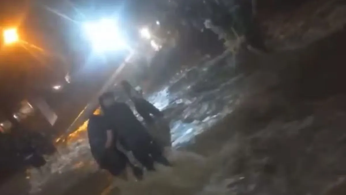 Pessoas atravessam rua tomada por enxurrada durante a noite, com água barrenta e forte correnteza, em meio à iluminação pública.