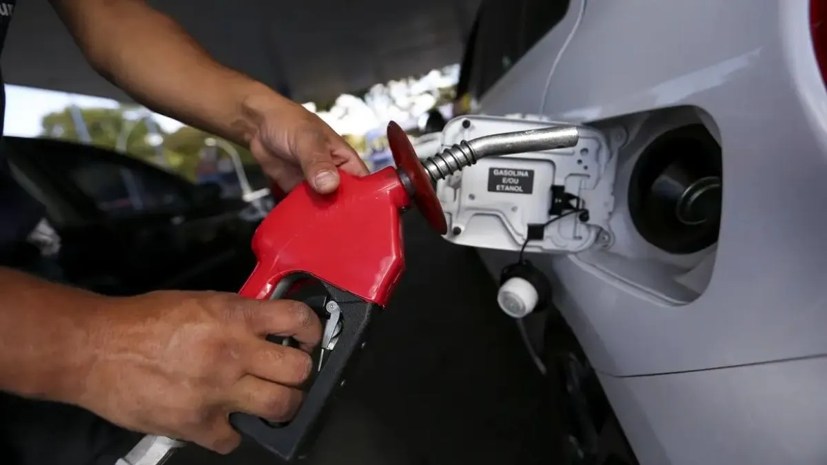 Frentista abastece carro branco com bomba de combustível vermelha em posto de gasolina, em meio à preocupação com possíveis aumentos nos preços.