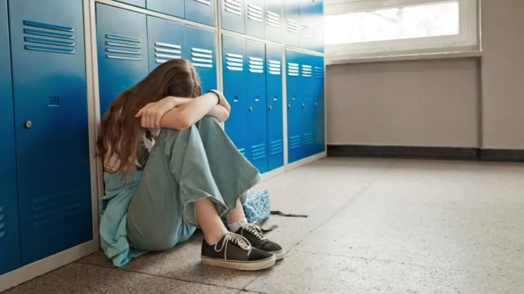 Dia Mundial de Combate ao Bullying: 27% dos adolescentes relatam agressões; veja os sinais de alerta 2 Menina sentada no chão de corredor escolar cobre o rosto com as mãos enquanto colegas riem ao fundo, em cena que representa o bullying na escola.