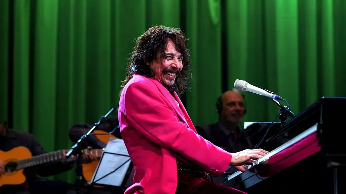 Músico de cabelos compridos e paletó rosa toca teclado durante apresentação, diante de cortinas verdes.