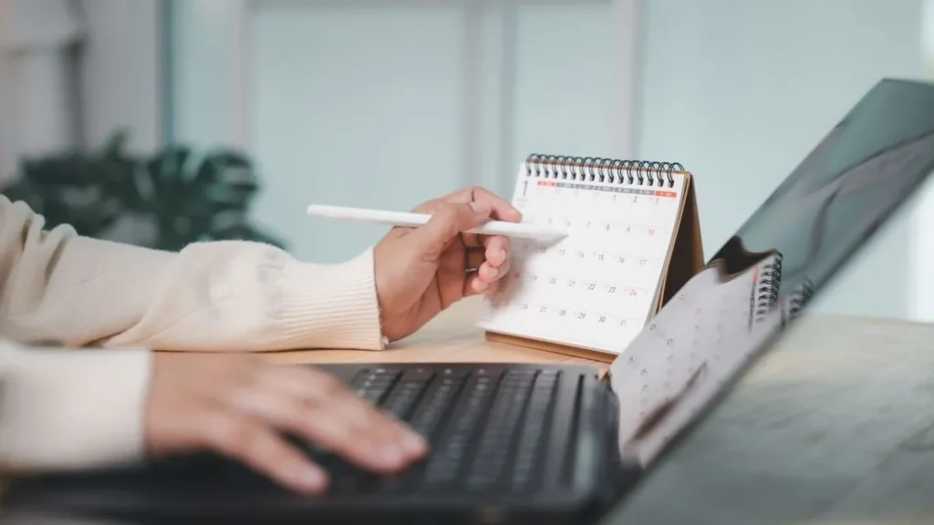 Pessoa utiliza notebook enquanto consulta calendário de mesa e organiza sua agenda de trabalho, ilustrando o planejamento das jornadas e folgas na escala 5x2.