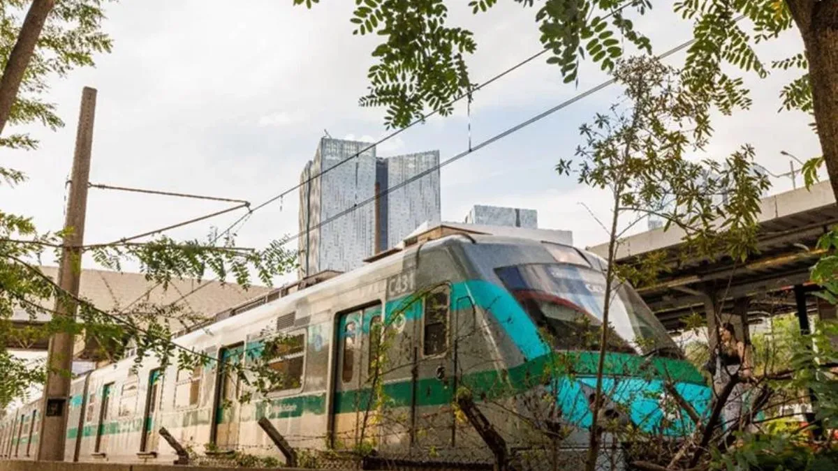 Trem nas cores prata e verde, próximo a uma plataforma, com árvores, fios elétricos e prédios ao fundo.
