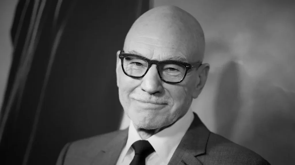 Retrato em preto e branco do ator britânico Patrick Stewart, de óculos, terno e gravata, com um leve sorriso diante da câmera.