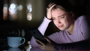 Jovem angustiada olha para o celular em ambiente escuro, representando o sofrimento emocional que pode ser provocado pelo cyberbullying nas redes sociais.