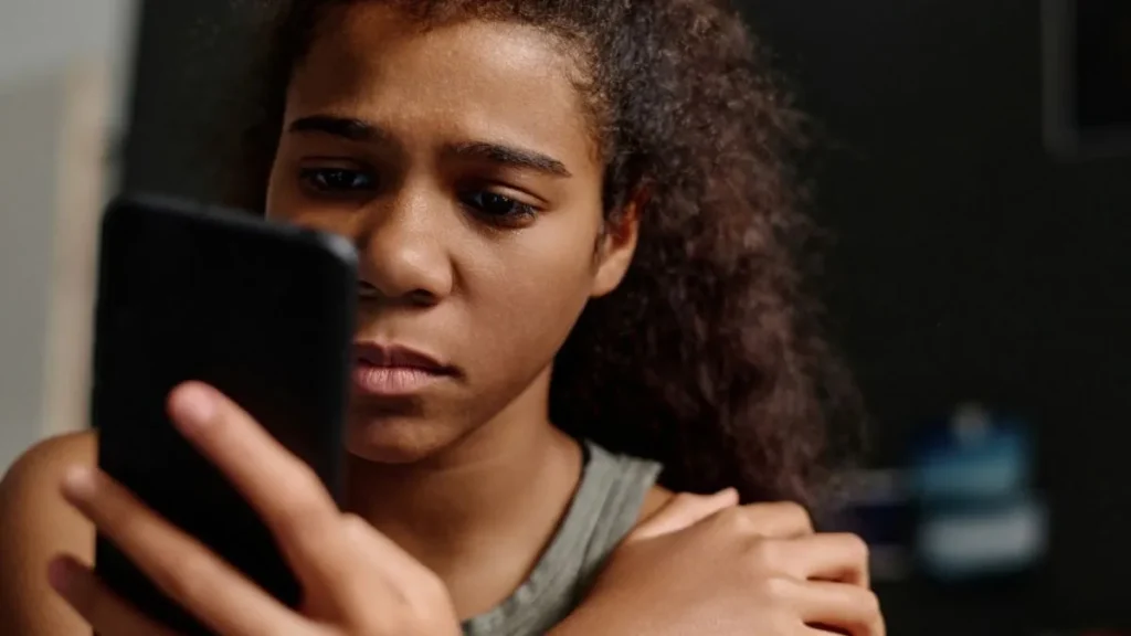 O que é cyberbullying? Conheça 6 formas de violência digital que podem passar despercebidas 3 Adolescente com expressão preocupada olha para o celular, em imagem que representa o sofrimento emocional causado pelo cyberbullying nas redes sociais.